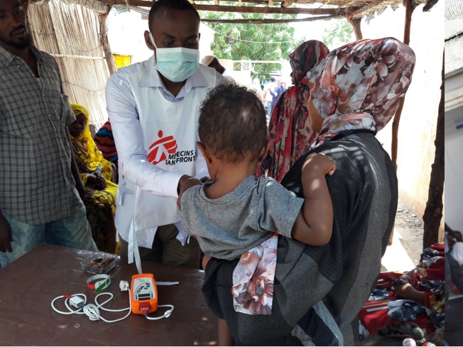 An MSF medical staff member examines a young child held by their caregiver during an outdoor consultation